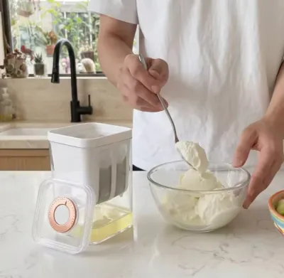A person spooning thick white yogurt into a glass bowl next to a compact Greek Yogurt Strainer on a counter.