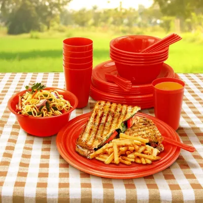 Red plastic picnic dinnerware set serving grilled sandwiches and noodles outdoors on a brown checkered tablecloth.